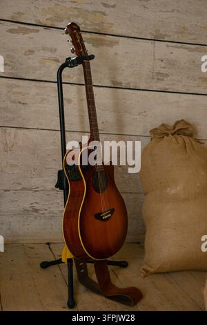 Riga, Lettonia - 4 ottobre 2026: Chitarra elettrica acustica esposta su un supporto accanto a un sacco di iuta in un fienile di legno stagionato, creando un'atmosfera rustica calda Foto Stock