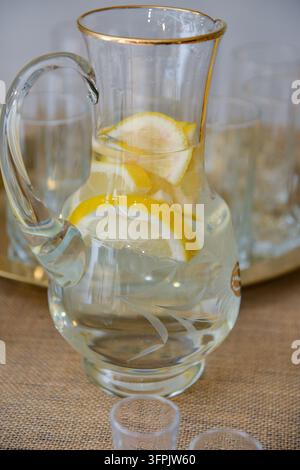Brocca di vetro trasparente riempita con acqua fresca e fette di limone su un tavolo rustico, creando un concetto di bevanda luminosa e rinfrescante per l'ospitalità, il benessere Foto Stock