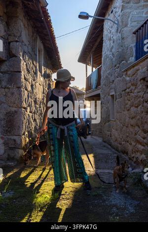 Donna incinta che passeggia con due cani al guinzaglio, godendosi una passeggiata serale lungo una stretta via di pietra in un villaggio tradizionale Foto Stock