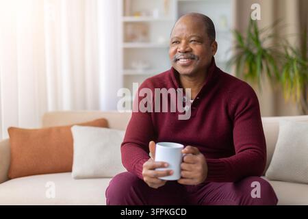 Uomo anziano nero sorridente seduto sul divano a casa e che tiene una tazza Foto Stock