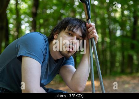 Un giovane si appoggia su bastoncini da trekking mentre riposa in una foresta verde. Foto Stock