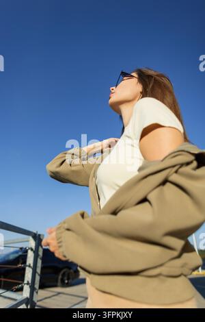 Scatto in basso di una giovane donna elegante con una giacca olivastra che le cade sulla spalla, che indossa occhiali da sole e una maglietta bianca contro un cielo blu chiaro o Foto Stock