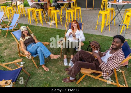Tre amici sorridenti e diversificati che si rilassano su sedie a sdraio sull'erba, godendosi bevande e conversazioni in un caffè all'aperto a Milano, Italia, celebrando gli amici Foto Stock