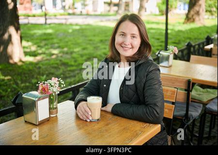 Donna sorridente seduta al tavolino da caffe' all'aperto con un bicchiere di latte, godendosi momenti tranquilli nell'ambiente del parco, luce naturale, ritratto di stile informale Foto Stock