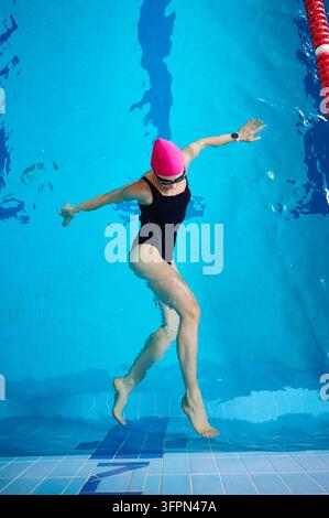 Giovane nuotatrice sott'acqua nella piscina, muovendo braccia e gambe, indossa costume da bagno, cuffia e occhialini, pratica allenamento acquatico e esercizi di fitness Foto Stock