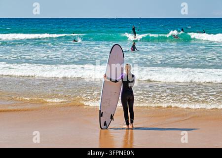 Vista posteriore di una donna in muta che tiene una tavola soft-top sul bordo dell'acqua mentre altri surfisti cavalcano le onde, Australia Foto Stock