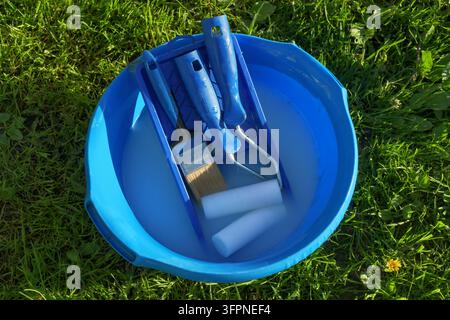 Rulli per vernice e pennello appoggiati in un bacino blu con acqua saponata sull'erba verde, che illustra l'ultima fase della pulizia degli strumenti per la pittura a base d'acqua. Foto Stock