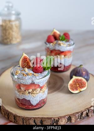 Dessert a strati servito in un bicchiere con fichi, lamponi, semi di chia e granola, uno strato bianco cremoso e guarnizione di menta fresca. Foto Stock