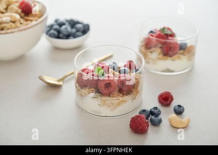 Dessert allo yogurt con granola e lamponi e mirtilli freschi in un bicchiere, servito con un cucchiaio, noci e bacche su una superficie chiara, con un altro y Foto Stock