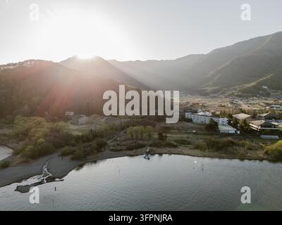 Vista aerea di un lago sereno circondato da montagne lussureggianti e edifici durante il tramonto a Narusawa, Yamanashi, Giappone. Foto Stock