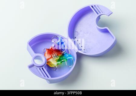 Il retain ortodontico arcobaleno riposa in una custodia ventilata viola aperta su uno sfondo pulito, una soluzione ordinata per riporre tra le sessioni di uso. Foto Stock