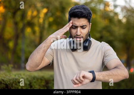 Uomo con cuffie e uno smartwatch, che controlla l'ora con un'espressione preoccupata, si sente sotto pressione e in ritardo mentre fa esercizio in un parco all'aperto Foto Stock
