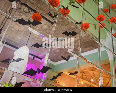 Decorazioni a pipistrello di Halloween su parete specchiata con riflessi di zucche a maglia arancioni, luci fantasma sospese. Le sagome di pipistrelli di Halloween creano un'atmosfera festosa Foto Stock
