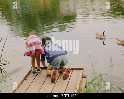 Madre e figlia in ginocchio su un molo di legno che osservano oche su un lago, Inghilterra, Regno Unito Foto Stock