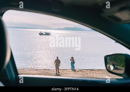 Uomo e donna che camminano verso la riva visti attraverso il finestrino dell'auto con mare scintillante e barca da pesca, Isola di Cai Chien, Quang Ninh, Vietnam Foto Stock