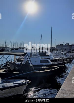 Croazia Piccoli motoscafi e barche a vela ormeggiati lungo un molo di cemento in una marina soleggiata con il sole splendente in alto nel cielo blu terso, forti riflessi. Foto Stock