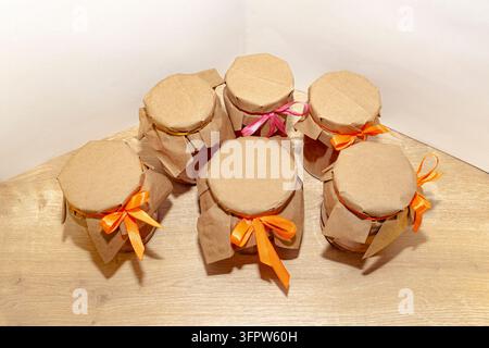 Marmellata di frutta fatta in casa in barattoli di vetro con coperchi di carta kraft e nastri colorati, disposti su un tavolo di legno per regali di cibo fatti a mano e occasioni festive Foto Stock