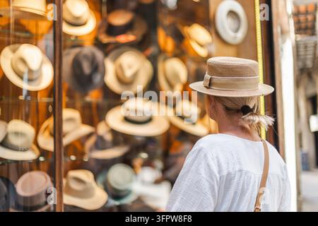 Donna con cappello di paglia osserva una colorata esposizione di cappelli al mercato La Boqueria di Barcellona, in Spagna. Foto Stock
