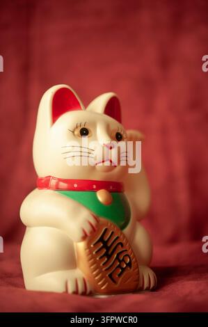 Gatto portafortuna giapponese Maneki-Neko bianco con una zampa alzata, moneta d'oro e bavero verde, posato su uno sfondo di tessuto rosso intenso Foto Stock