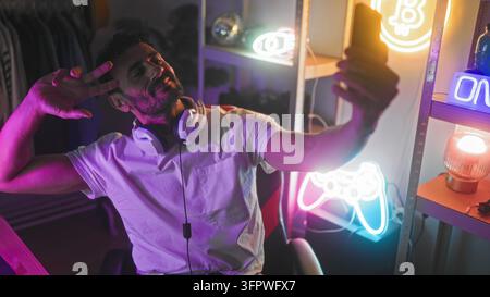 Uomo con auricolari e maglietta bianca che fa il segno della pace e tiene uno smartphone per un selfie tra luci al neon e un cartello "On Air" in studio; connessione giocosa. Foto Stock