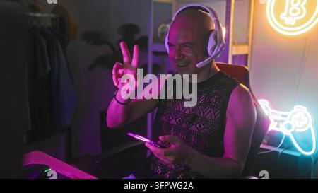 Uomo con auricolari tiene uno smartphone con entrambe le mani, sorridendo e toccando lo schermo mentre è seduto a una scrivania da gioco in uno studio al neon; concentrazione gioiosa. Foto Stock