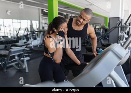 Uomo nero preoccupato che consola una donna che avverte disagio durante l'esercizio sul tapis roulant in palestra Foto Stock