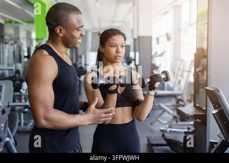 Personal trainer nero muscoloso che guida una donna atletica nell'esecuzione di curl bicipiti con i manubri Foto Stock