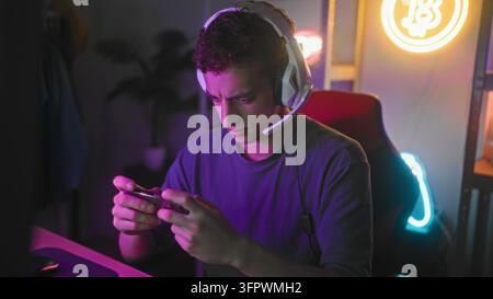 Giovane uomo con auricolari tiene uno smartphone e agita la mano mentre è seduto in un setup di gioco da camera da letto; gioia amichevole nel gaming. Foto Stock
