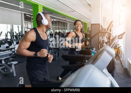 Coppia nera atletica e felice che fa jogging su tapis roulant adiacenti in un moderno centro fitness Foto Stock