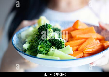 Immagine ritagliata di una giovane donna in maglietta bianca che mostra broccoli e carote in un piatto alla telecamera. Concetto di alimentazione sana. Foto Stock