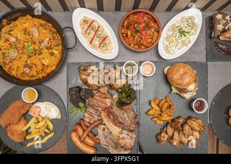 Banchetto spagnolo visto dall'alto con paella, carni grigliate miste, pollo arrosto, cotoletta impanata con uovo, burger, alette, wedges, insalate di patate e ratatouille Foto Stock