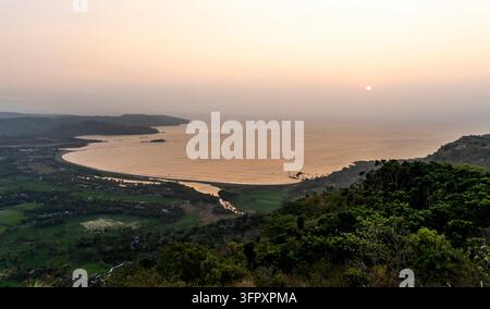 Tramonto pacifico su una baia costiera curva, con mare calmo, colline verdi, case di villaggio sparse e il sole che splende sopra l'orizzonte. Foto Stock