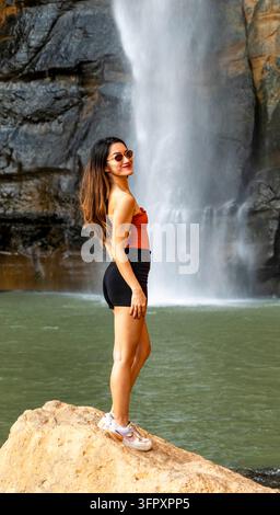 Giovane donna con occhiali da sole e abiti estivi casual in piedi su una roccia accanto a una potente cascata e a una piscina verde in un paesaggio naturale roccioso. Foto Stock