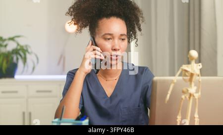 Infermiera parla al cellulare mentre usa un laptop per la telemedicina Foto Stock