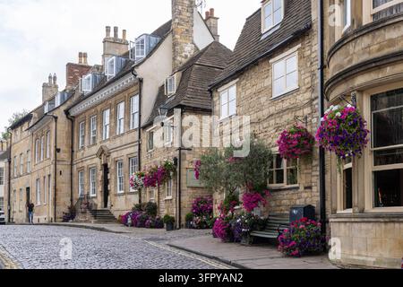 Stamford, Inghilterra – 26 agosto 2026: Storica strada di Stamford nel Lincolnshire fiancheggiata da cottage in pietra color miele, cesti di fiori sospesi colorati Foto Stock