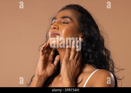 Donna indiana in posa, tocca il collo e la mascella in studio con illuminazione soffusa, indossa un top a spaghettini bianco. Ritratto, sereno, calmo, caldo, beige, minimale, elegante Foto Stock