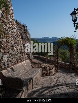 Una panca di pietra invecchiata offre un punto di riposo su un sentiero in pietra terrazzato, offrendo una vista panoramica sulle montagne lontane sotto un cielo azzurro a Mallorc Foto Stock