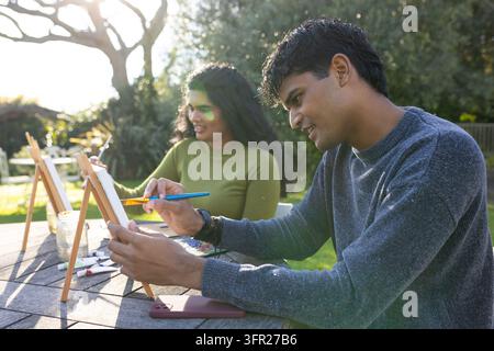 Coppia indiana in maglioni che dipinge su piccole tele a un tavolo da picnic nel cortile con pennelli Foto Stock