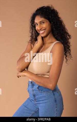 Donna afroamericana che sorride e posa davanti a uno sfondo studio marrone chiaro indossando una canottiera beige e jeans. Ritratto, sicura di sé, riccia, calda, morbida, illuminazione Foto Stock