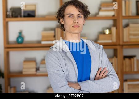 Uomo adulto in piedi a casa con una felpa blu con scollo a T-shirt, con libri e un vaso turchese sullo scaffale Foto Stock