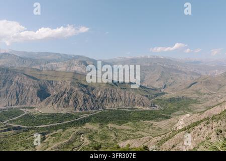 Vista ad angolazione elevata di ampie catene montuose aride e di una valle fluviale verde sotto un cielo estivo azzurro, che mostra canyon stratificati drammatici e una geologia accidentata Foto Stock