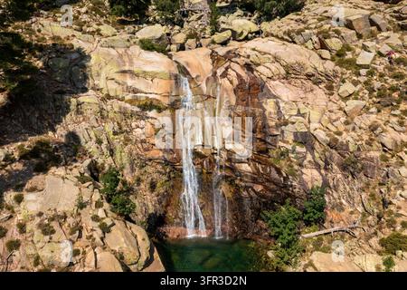 Una cascata di montagna scorre su una scogliera rocciosa in un piccolo bacino circondato da massi di granito e vegetazione rada alla luce del giorno. Foto Stock