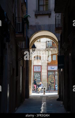 RIPOSO ILLUMINATO ALLA FINE DI UN VICOLO SCURO, REPORTAGE FOTOGRAFICO DI STRADA A BARCELLONA, BARCELLONA, SPAGNA, 2019: Incorniciati dalle ombre profonde di un vicolo stretto e da un arco, due persone sono sedute su sedie rilassandosi in una piccola macchia di luce solare brillante. INFO: Barcellona è una vivace città costiera spagnola rinomata per la sua architettura iconica, le spiagge mediterranee, il quartiere gotico storico, la ricca cultura catalana, il calcio di classe mondiale e un'atmosfera metropolitana vivace piena di arte, cibo e musica. Foto Stock