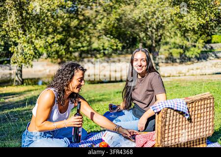 Due donne sorridenti si godono vino e snack su una coperta da picnic in un parco verde e soleggiato, con un cestino di vimini e alberi dietro di loro. Foto Stock