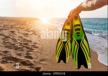 Concetto di viaggio estivo, sport e tempo libero. Mano maschile che tiene l'attrezzatura per lo snorkeling o le immersioni subacquee sullo sfondo della spiaggia e del sole Foto Stock