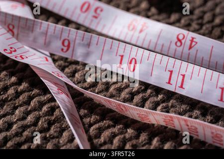 Nastro metrico bianco con numeri rossi appoggiato su una superficie lavorata a maglia marrone strutturata, indicante misurazione e precisione Foto Stock