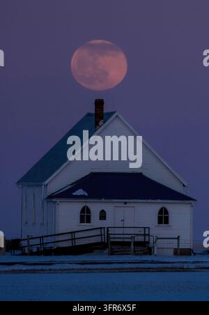 Grande luna piena che sorge su una piccola chiesa bianca in un paesaggio innevato Foto Stock