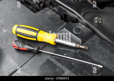 Un grande cacciavite giallo intercambiabile e un cacciavite nero sottile riposano insieme su un pannello del vano motore in plastica nera. La vista ravvicinata evidenzia Foto Stock