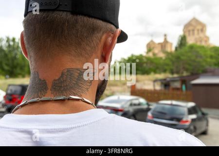 Un uomo con ali tatuate sul collo si trova con la Cattedrale di San Gregorio Illuminatore sullo sfondo a Yerevan, Yerevan, Armenia Foto Stock