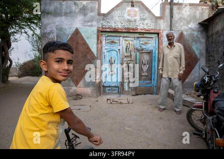 Ragazzo sorridente in bicicletta davanti a una porta blu consumata mentre un anziano osserva, Gujarat, Mandvi, India Foto Stock
